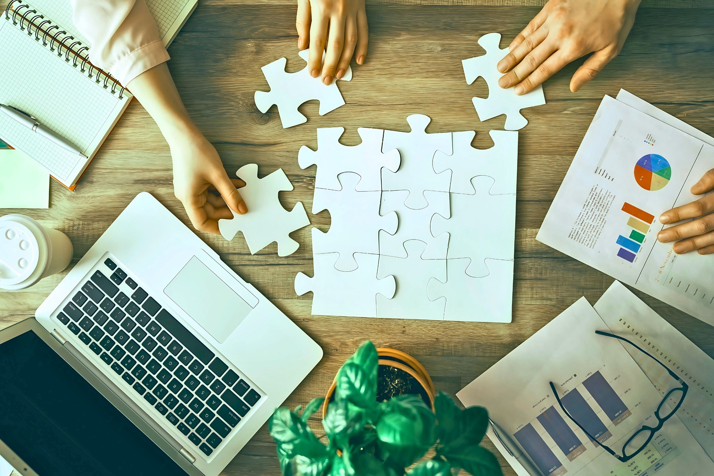 Hands assembling puzzle pieces on a table with charts and laptop, symbolizing teamwork in content strategy